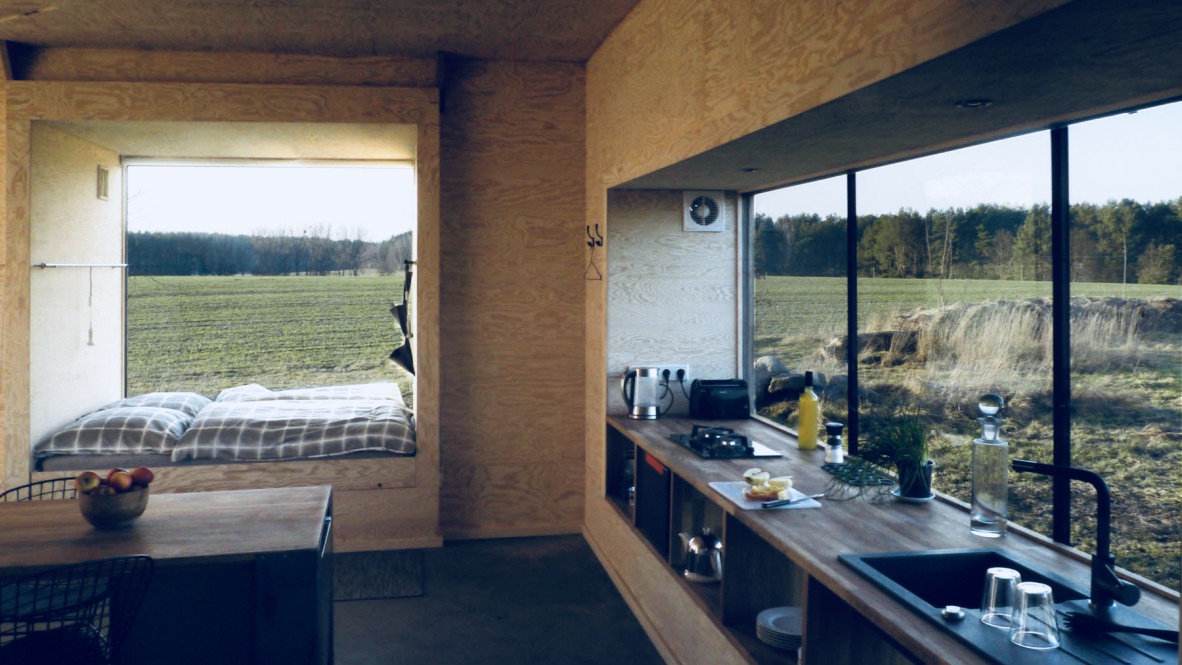 Das Bild zeigt ein Minihaus von innen. Im Minihaus sieht man eine Küche mit einem großen Fenster und einem Schlafplatz.